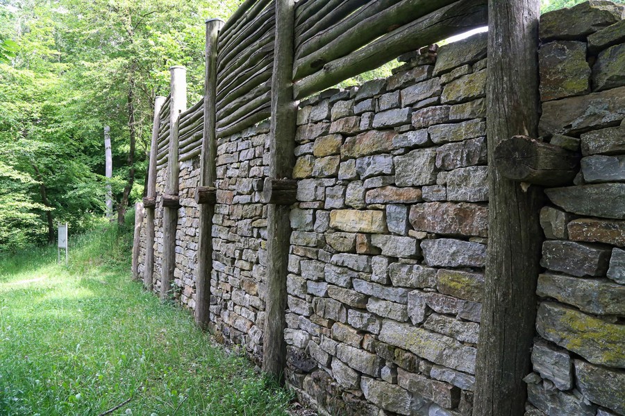 Eine hohe Steinmauer steht auf einer Wiese. Über der Mauer wurden noch mit Holzbalken die Mauer erhöht.