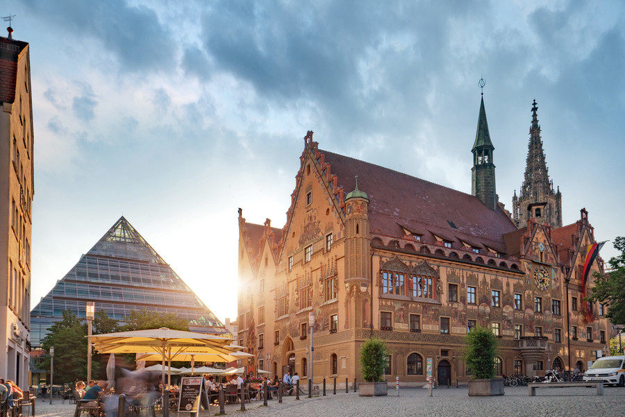 Blick auf das historische Rathaus in Ulm. Davor ist der Marktplatz mit einer Gaststätte. Die Menschen sitzen unter gelben Sonnenschirmen.