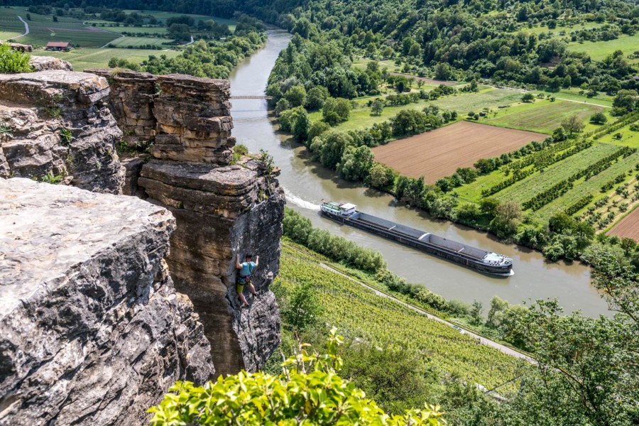 Ein Kletterer klettert an einem steilen Felsen. Im Hintergrund fährt ein großes Schiff auf einem Fluss.