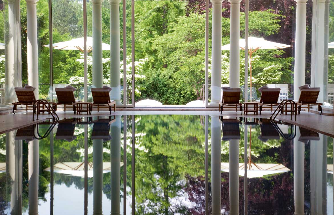 In der Mitte ist ein großer Pool. Dahinter stehen sechs Liegestühle und viele weiße Säulen. Im Hintergrund stehen Bäume und Büsche.