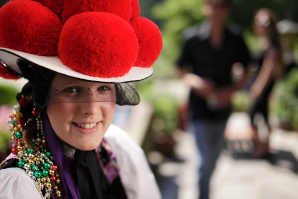 Ein Mädchen trägt eine Tracht und einen Schwarzwälder Bollenhut mit roten runden Bollen darauf. Sie lacht in die Kamera. 