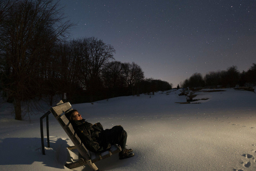 Eine Person sitzt auf einer Holzbank bei Dunkelheit in der verschneiten Landschaft und schaut nach oben in den Sternenhimmel.