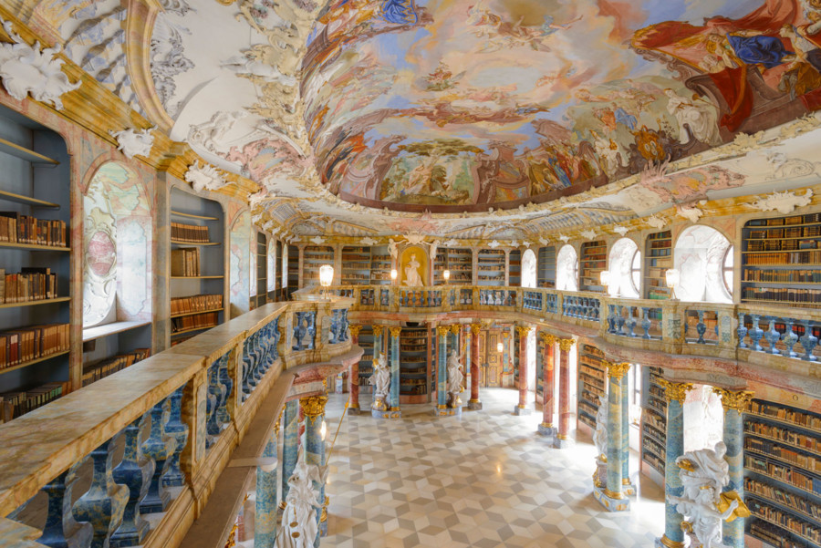 Innenaufnahme des Bibliothekssaal des Klosters. Die Decke ist verziert mit Malereien. An den Wänden sind große Regale mit vielen Büchern.