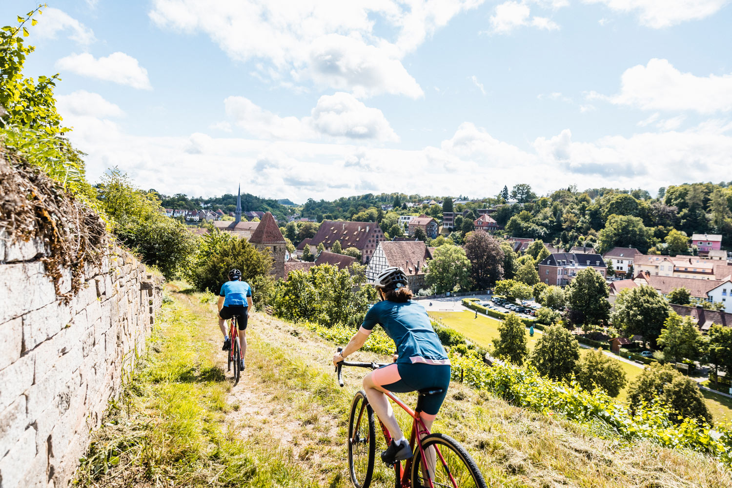 Ein Radfahrer und eine Radfahrerin fahren durch die Weinberge auf das Kloster Maulbronn zu. Der Himmel ist leicht bewölkt.