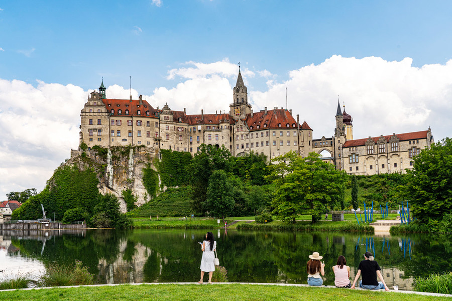 Blick auf das Hohenzollernschloss Sigmaringen. Vor dem Schloss ist ein großer See an dem Menschen sitzen.