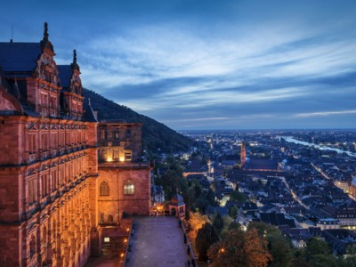 Blick von oben auf die beleuctete Fassade von Schloss Heidelberg und die Stadt Heidelberg bei Dämmerung. Der Himmel ist Wolkenverhangen. 