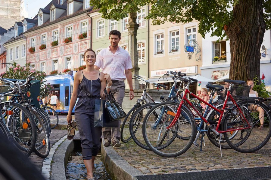 Ein Mann und eine Frau laufen barfuß durch die berühmten Bächle in Freiburg in der Altstadt. Links und rechts von ihnen stehen Fahrräder. Hinter ihnen stehen Häuser.