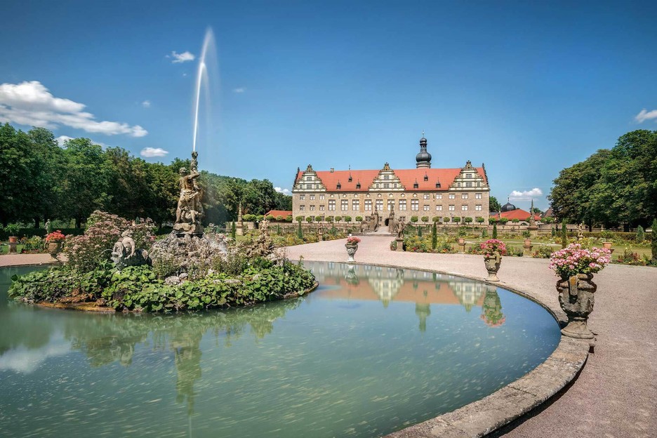Schloss und Schlossgarten Weikersheim. Im Vordergrund ist ein großer Brunnen mit einer Statue aus der Wasser fließt. Im Hintergrund steht das Schloss.